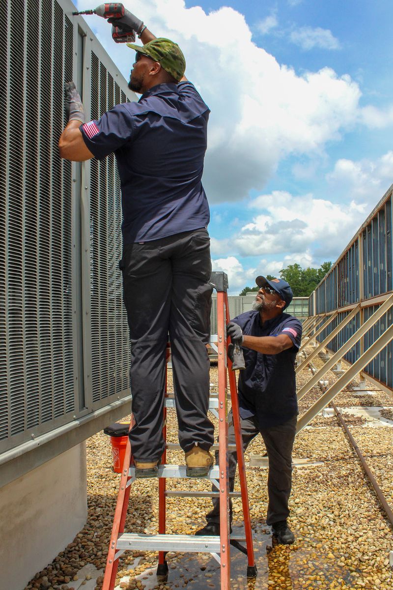 Two Willis Mechanical technicians on a rooftop fastening a louvered panel onto a large condensing unit under a blue sky